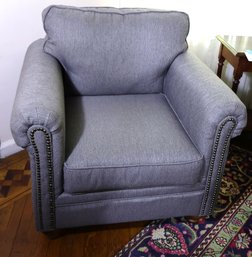 Cozy Club Chair With Nail Head Trim On Rolled Arms And Bun Feet Upholstered In Textured Grayish Blue Fabric.