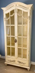 A Distressed Cream-colored Display Cabinet With Ornate Carvings And Glass Doors