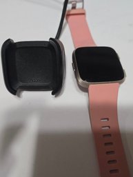 A Fitbit Versa Series Smartwatch In A Rose Gold/peach Color With Charger