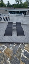 Set Of Two Black Woven Outdoor Chaise Lounge Chairs With A Small Matching Side Table.